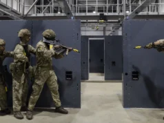 Five soldiers in camouflage gear practice room-clearing behind gray modular walls in an indoor training facility.
