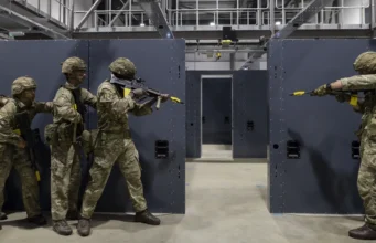 Five soldiers in camouflage gear practice room-clearing behind gray modular walls in an indoor training facility.