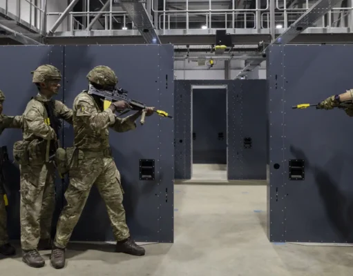 Five soldiers in camouflage gear practice room-clearing behind gray modular walls in an indoor training facility.