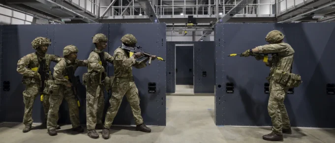 Five soldiers in camouflage gear practice room-clearing behind gray modular walls in an indoor training facility.