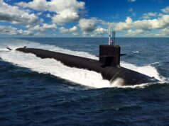 America places huge order for 14 more nuclear submarines A dark submarine gliding at the ocean surface, creating a white wake with a blue sky and clouds above, a small American flag on the conning tower.