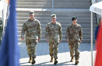 European officer to lead Ukraine aid command within a year Three soldiers in camouflage walk down a wide set of stairs, flags blurred in the foreground.