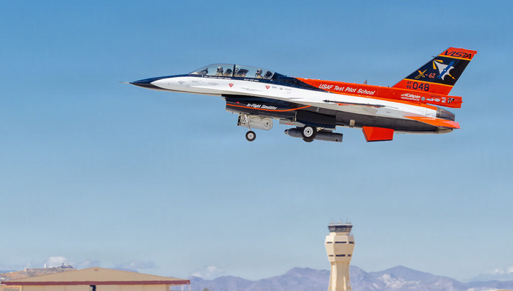 USAF Test Pilot School jet trainer with two crew taking off from an airbase; control tower in the background.