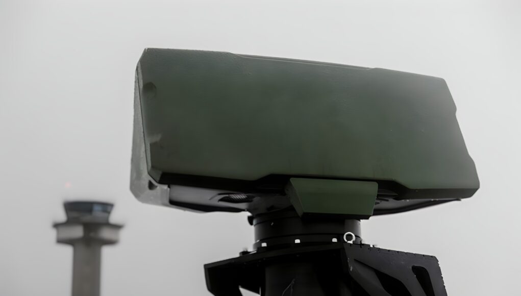 grifaffe33.remini-enhanced Close-up of a green military weapon mount on a vehicle with a blurred distant tower in the background