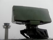 Close-up of a green military weapon mount on a vehicle with a blurred distant tower in the background