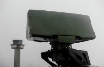 Close-up of a green military weapon mount on a vehicle with a blurred distant tower in the background