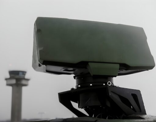Close-up of a green military weapon mount on a vehicle with a blurred distant tower in the background