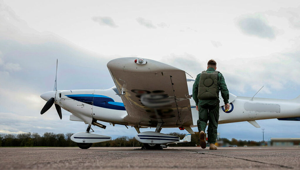 grobviking Pilot in a green flight suit walks toward a small white-and-blue propeller plane on a runway.