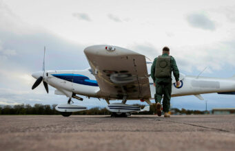 Air cadet gliders to serve to mid-century after upgrade Pilot in a green flight suit walks toward a small white-and-blue propeller plane on a runway.