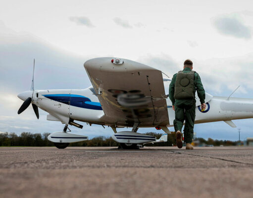 Pilot in a green flight suit walks toward a small white-and-blue propeller plane on a runway.
