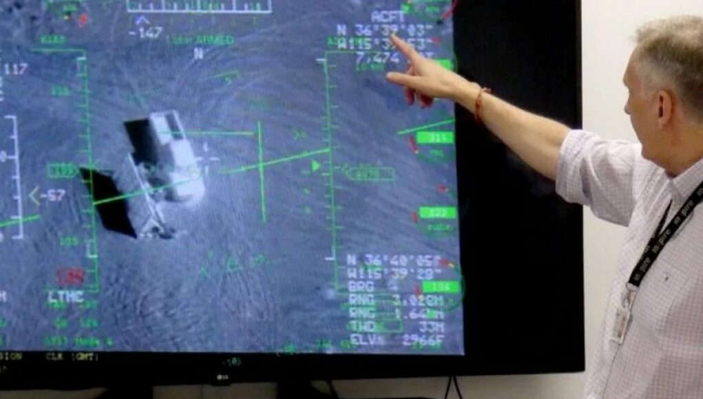 Person in a light shirt points at a large screen displaying a flight navigation/avionics interface with green markers and data.