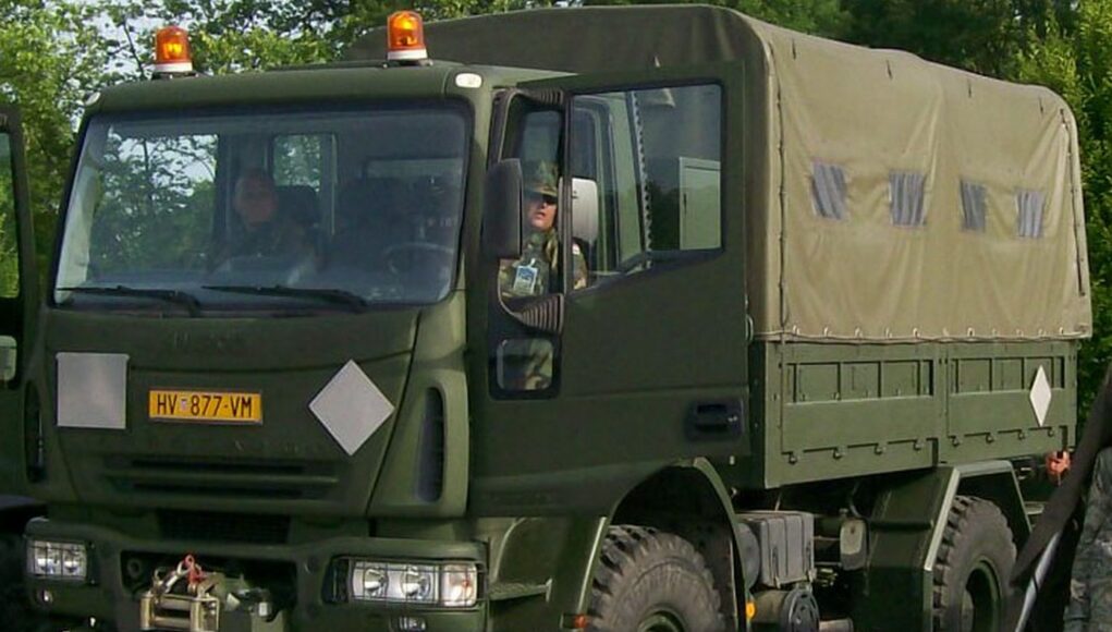 iveco Front three-quarter view of an olive-drab military cargo truck with a canvas-covered rear compartment driving on a road with amber warning lights overhead.