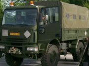 Front three-quarter view of an olive-drab military cargo truck with a canvas-covered rear compartment driving on a road with amber warning lights overhead.