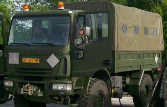Front three-quarter view of an olive-drab military cargo truck with a canvas-covered rear compartment driving on a road with amber warning lights overhead.