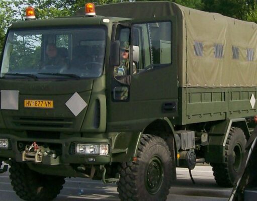 Front three-quarter view of an olive-drab military cargo truck with a canvas-covered rear compartment driving on a road with amber warning lights overhead.