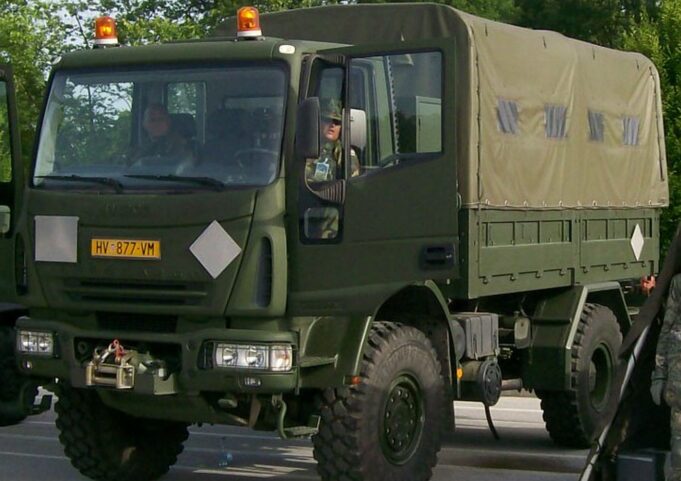 Front three-quarter view of an olive-drab military cargo truck with a canvas-covered rear compartment driving on a road with amber warning lights overhead.