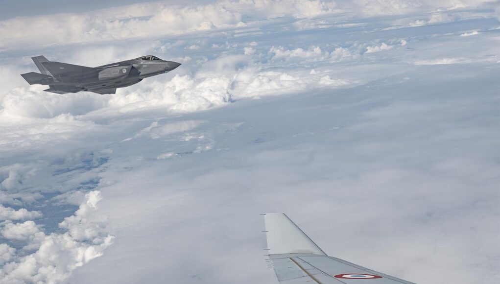 kZwmq1568 Military jet flying above clouds, seen from a passenger plane window; French Air Force markings visible on the fuselage.