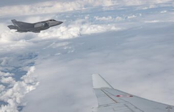 Military jet flying above clouds, seen from a passenger plane window; French Air Force markings visible on the fuselage.