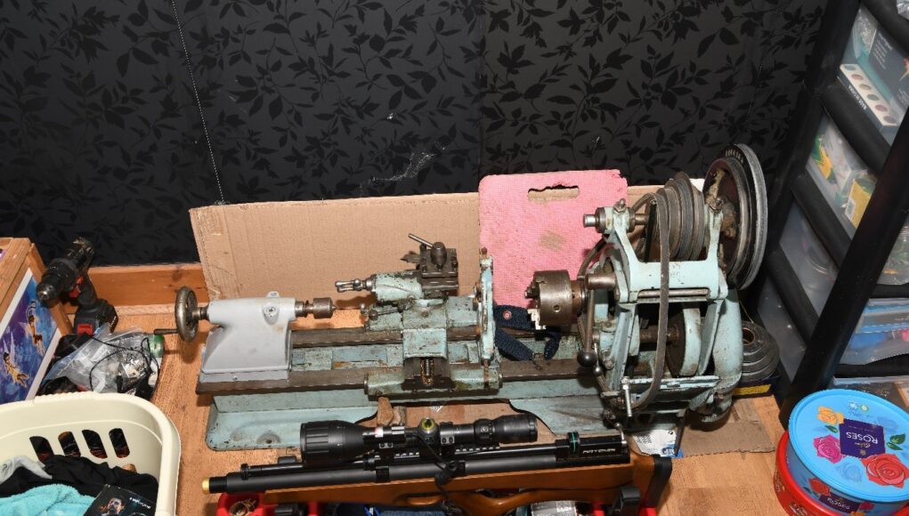 A small metal lathe on a workbench with a belt-driven headstock, tooling, and a pink foam block behind it; cluttered workshop scene around it.