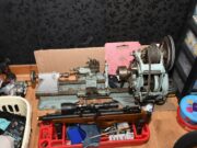 A small metal lathe on a workbench with a belt-driven headstock, tooling, and a pink foam block behind it; cluttered workshop scene around it.