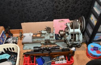 A small metal lathe on a workbench with a belt-driven headstock, tooling, and a pink foam block behind it; cluttered workshop scene around it.