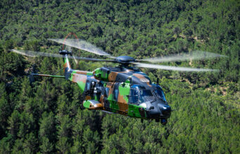 Camouflage military helicopter in flight above a dense green forest.