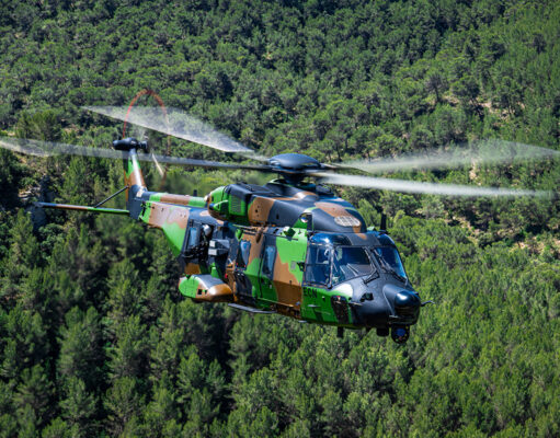 Camouflage military helicopter in flight above a dense green forest.
