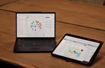 Laptop shows a circular data dashboard while a tablet displays a map dashboard on a wooden table.