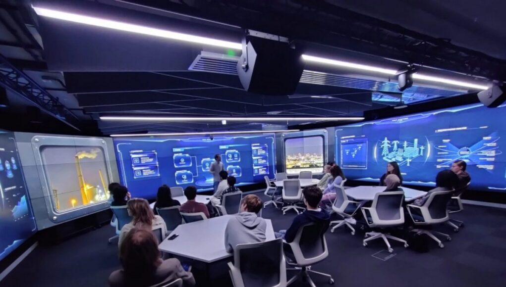 Group of people seated around a U-shaped table in a high-tech blue-glow control room, watching a presenter at the front.
