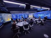 Group of people seated around a U-shaped table in a high-tech blue-glow control room, watching a presenter at the front.