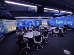 QinetiQ trains 200 students on NCSC cyber security course Group of people seated around a U-shaped table in a high-tech blue-glow control room, watching a presenter at the front.