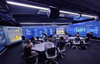 QinetiQ trains 200 students on NCSC cyber security course Group of people seated around a U-shaped table in a high-tech blue-glow control room, watching a presenter at the front.