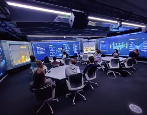 Group of people seated around a U-shaped table in a high-tech blue-glow control room, watching a presenter at the front.