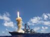 Military ship launches a rocket, bright flame and smoke plume rise from the deck into a blue sky over the ocean.
