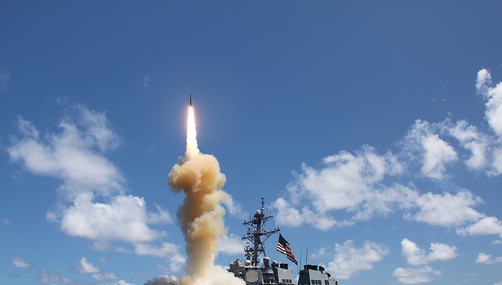 Military ship launches a rocket, bright flame and smoke plume rise from the deck into a blue sky over the ocean.