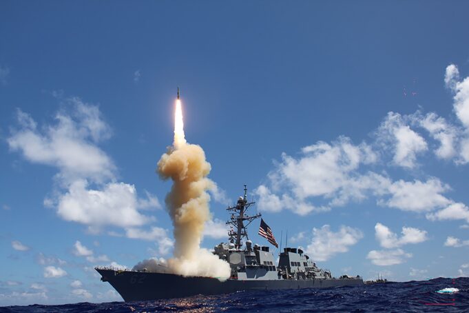 Military ship launches a rocket, bright flame and smoke plume rise from the deck into a blue sky over the ocean.
