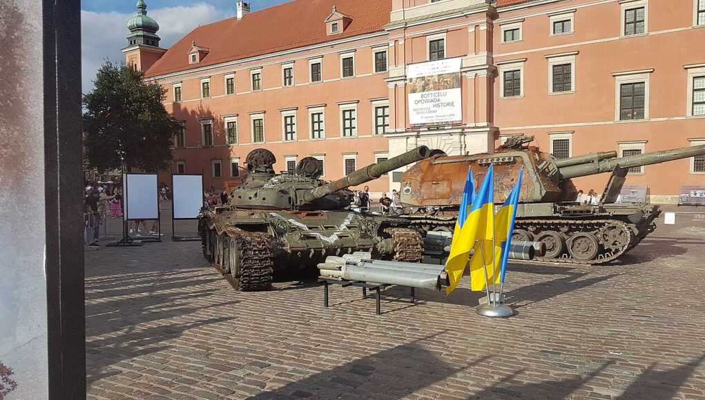 t72 Two military tanks on a cobblestone plaza in front of a peach-colored historic building; Ukrainian flags in the foreground.