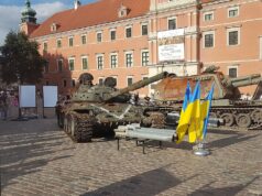 Two military tanks on a cobblestone plaza in front of a peach-colored historic building; Ukrainian flags in the foreground.