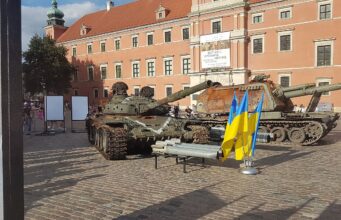 Two military tanks on a cobblestone plaza in front of a peach-colored historic building; Ukrainian flags in the foreground.