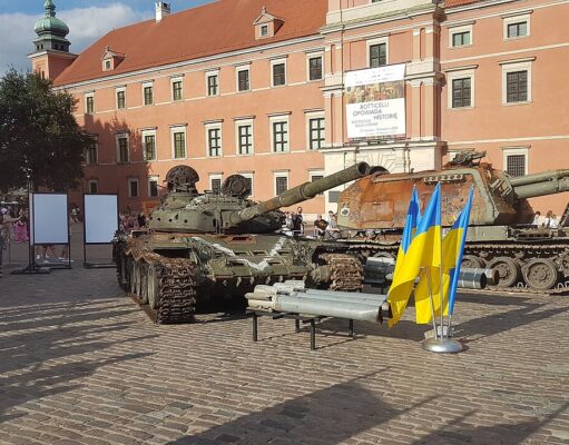 Two military tanks on a cobblestone plaza in front of a peach-colored historic building; Ukrainian flags in the foreground.