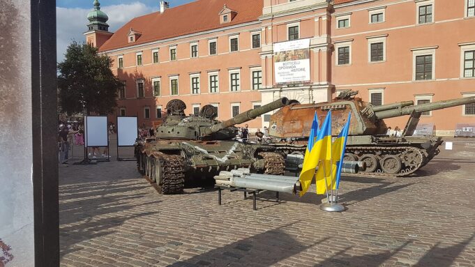 Two military tanks on a cobblestone plaza in front of a peach-colored historic building; Ukrainian flags in the foreground.