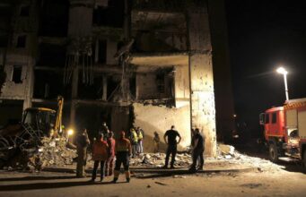 Nighttime scene of a collapsed building with rescue workers and a fire truck inspecting the rubble.