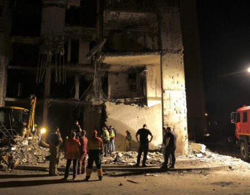 Nighttime scene of a collapsed building with rescue workers and a fire truck inspecting the rubble.