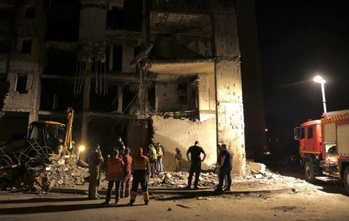 Nighttime scene of a collapsed building with rescue workers and a fire truck inspecting the rubble.